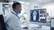 © javu - หมอกำลังนั่งอยู่ที่โต๊ะพร้อมคอมพิวเตอร์ หน้าจอที่ใหญ่กว่ากำลังแสดงภาพเอ็กซ์เรย์ หมอกำลังดูหน้าจอ