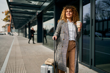 At the bus station, a businesswoman pushes her suitcase, coffee cup in hand.