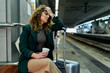 At the railway station, a tired woman sits, holding a suitcase, enduring a headache.