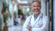 A successful respectable doctor in a white uniform and a stethoscope stands against the background of light walls in a modern clinic. Portrait of a handsome smiling doctor. Healthcare industry concept