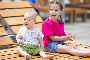 Wall Mural - Cute children little girl and newborn baby boy sitting on wooden beach chair near the sea during family vacation. Summer travel concept.