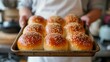 © Kate Mova - a man in a white shirt and apron holds a baking sheet with six freshly baked buns sprinkled with sesame seeds.
