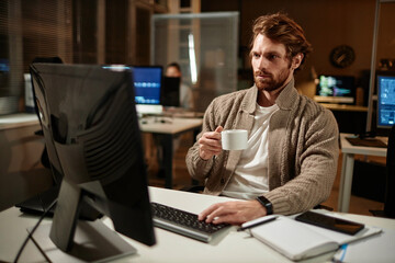 portrait of bearded man using computer in office and drinking coffee while working late at night, co