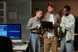 © Seventyfour - Waist up portrait of three young people using laptop standing in IT office at night, copy space