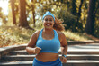 Happy smiling overweight woman jogging in park in summer. Portrait of cheerful beautiful fat plump chubby stout young lady in blue sports bra and sweatband running down stone steps in green city park