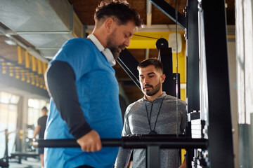 Wall Mural - Young coach assisting mature man in exercising in gym.