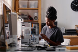 Fototapeta  - Black female technician sitting at table in workshop and repairing system unit using screwdriver