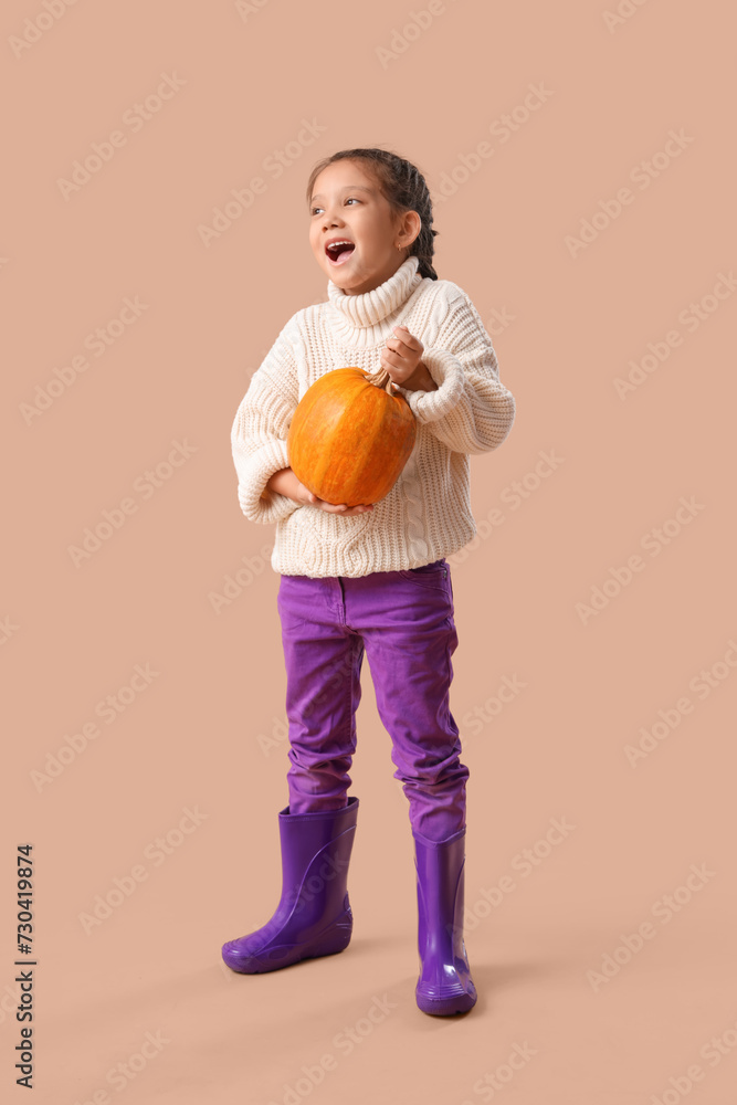 Little girl in rubber boots with pumpkin on beige background