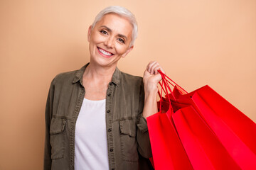 Sticker - Portrait of happy short blond hair lady wearing khaki shirt holding red bright packages purchases isolated on beige color background