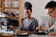 © AnnaStills - Young black woman sitting at workshop table and smiling at camera while repairing electronic device