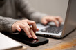 © bongkarn - A woman using her smartphone while typing on a keyboard, working on her laptop at a table.