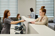 © pressmaster - Young female receptionist in grey uniform helping female client with medical document by administrator counter in clinics