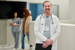 © pressmaster - Successful male clinician in labcoat looking at camera with smile while standing against reception counter and two young women