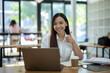 © Wasana - Cheerful young Asian businesswoman giving a thumbs up while working with a laptop at a wooden table in a bright office..
