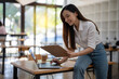 © Wasana - An energetic Asian businesswoman is actively working with a laptop while referencing notes on a clipboard in a vibrant office space..