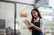 © Wasana - Smiling Asian cafe owner writes in her notebook, ready to welcome customers, with a 'Welcome Open' sign hanging in the background..