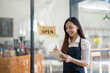 © Wasana - Smiling Asian cafe owner writes in her notebook, ready to welcome customers, with a 'Welcome Open' sign hanging in the background..