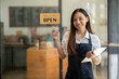 © Wasana - A friendly Asian shop owner smiles and points to a 'Welcome Open' sign, holding a tablet at the entrance of her cafe..