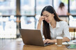 © Wasana - A stressed young Asian businesswoman massages temples while focusing on a laptop screen in a well-lit office environment..