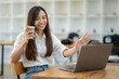 © Wasana - A friendly Asian businesswoman waves hello during a video call on her laptop, holding a coffee cup in a bright office setting..