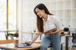 © Wasana - An energetic Asian businesswoman is actively working with a laptop while referencing notes on a clipboard in a vibrant office space..
