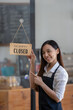 © Wasana - Asian female cafe owner with a welcoming smile presents the open sign at her shop, signaling readiness for customers..