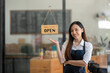 © Wasana - Asian female cafe owner with a welcoming smile presents the open sign at her shop, signaling readiness for customers..