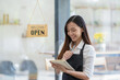 © Wasana - Smiling Asian cafe owner writes in her notebook, ready to welcome customers, with a 'Welcome Open' sign hanging in the background..
