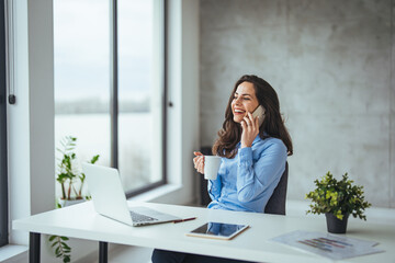 Wall Mural - Young businesswoman sitting at her desk talking on phone and working on computer. Female executive working in modern workplace. Young businesswoman using mobile phone and laptop