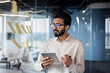 © Tetiana - Serious and focused young Indian man working in office with tablet, standing near glass board with notes, processing data, thinking about project