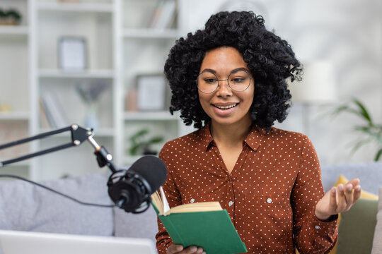 Home recording online studio. Young smiling African American woman sitting on sofa in front of microphone and laptop and reading book, falling asleep to podcast, giving sermon