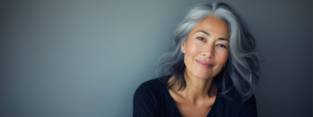 Happy smiling stylish confident 50 years old Asian female professional standing looking at camera at gray background. Portrait of sophisticated grey hair woman advertising products.