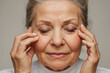 Senior caucasian woman with closed eyes holding fingers on skin near eyes to show eyes aging and care