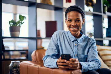 Wall Mural - Portrait of a smiling African woman, holding a mobile phone, sitting on the sofa at the office.