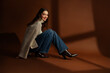 Fashionable happy smiling woman wearing oversized white sweater, wide leg jeans, leather cowboy boots, posing on brown background. Full-length studio fashion portrait. Copy, empty blank space for text