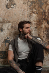 Wall Mural - beautiful brutal hipster man with a beard and hairstyle with a tattoo in a white T-shirt with a sweater and jeans sits on the floor near a concrete grunge texture wall