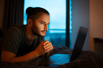 Wall Mural - Serious young guy lying on bed and working home on laptop. Looking at the screen.