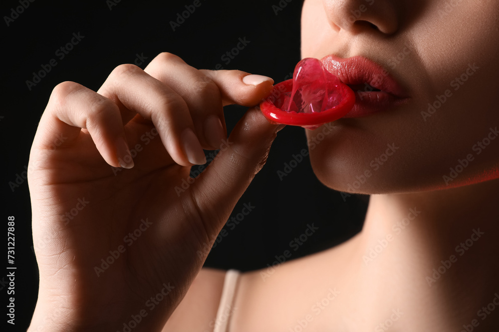 Young woman with condom on dark background, closeup. Sex concept