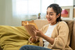 © Chanakon - Asian beautiful woman sitting reading book on cozey couch sofa in living room with sun light morning. People female stay home part time relax at home.