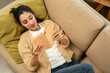 © Chanakon - Asian beautiful woman lay down on cozey couch sofa reading book in living room with sun light morning. People female stay home part time relax at home.
