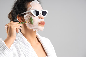 Wall Mural - Young African-American woman in sunglasses with sheet mask massaging face on light background, closeup