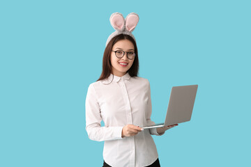 Poster - Young Asian businesswoman in bunny ears with laptop on blue background. Easter celebration