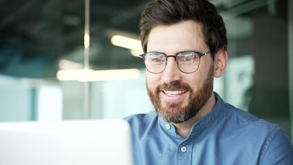 Happy smiling handsome businessman working on a laptop sitting at a workplace in business office. A satisfied bearded entrepreneur in glasses chats online, read the good news on the computer. Close up