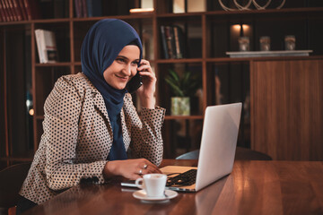 Wall Mural - Successful smiling Arab woman in hijab working inside modern office, business woman using a smart phone Muslim woman using laptop at work