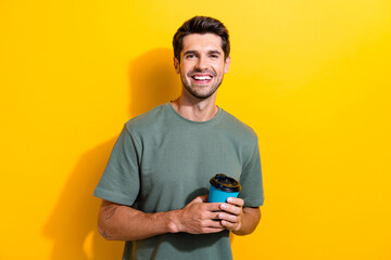 Poster - Portrait of toothy beaming handsome guy with stubble holding takeaway cup of coffee in morning isolated on yellow color background