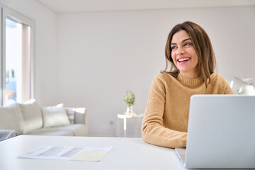 Happy smiling older mature middle aged woman using laptop computer sitting at table in living room, hybrid working, elearning online with technology device at home, looking away at copy space.