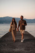 Happy couple strolling on asphalt pier at shore of Adriatic sea and enjoying sunset. Smiling husband and wife in evening outfits walking hand in hand, talking and smiling.