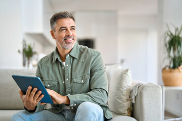 Happy middle aged man using digital tablet relaxing on couch at home. Mature male user holding tab computer holding pad technology device sitting on sofa in living room looking away. Copy space.
