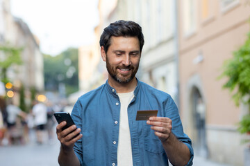 Mature bearded man with credit card and smartphone on city street in the evening