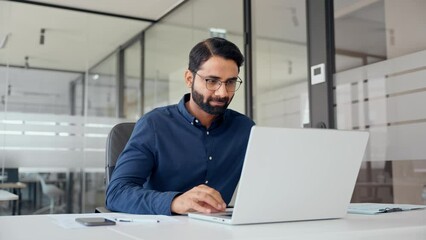 Smiling Indian business man looking at laptop using computer working in office. Busy professional businessman employee or company executive searching online solutions at workplace.
